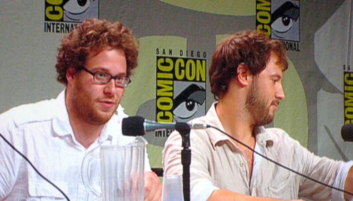 Seth Rogen in glasses and a white shirt beside Evan Goldberg at a panel table with microphones
