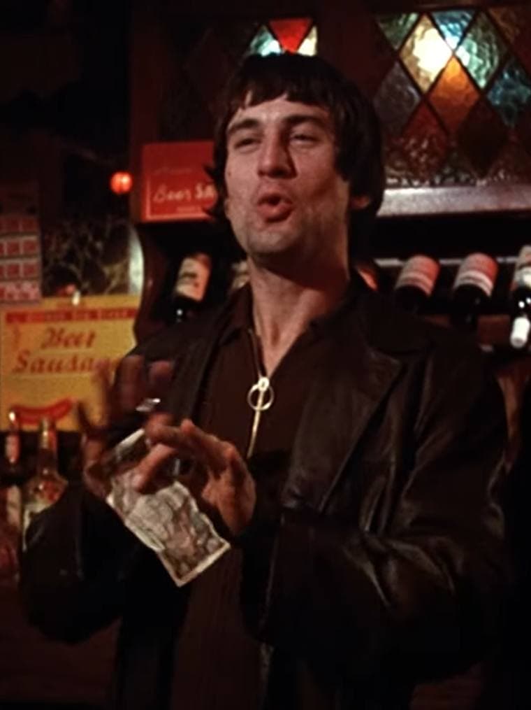 A young man with dark tousled hair and a leather jacket talks in a dim bar, holding a glass