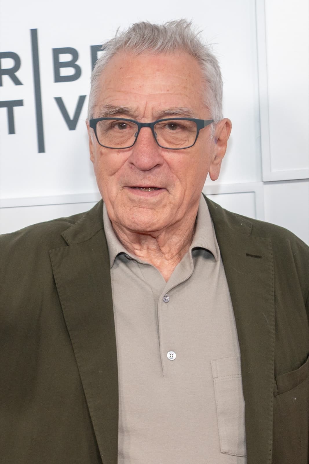 Robert De Niro, grey-haired with glasses, in an olive jacket in front of a white festival backdrop