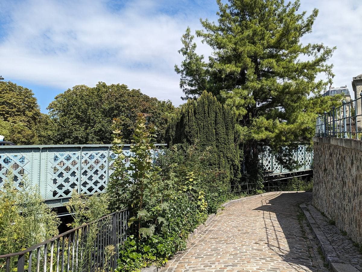 An iron road bridge over a leafy cemetery with a paved ramp