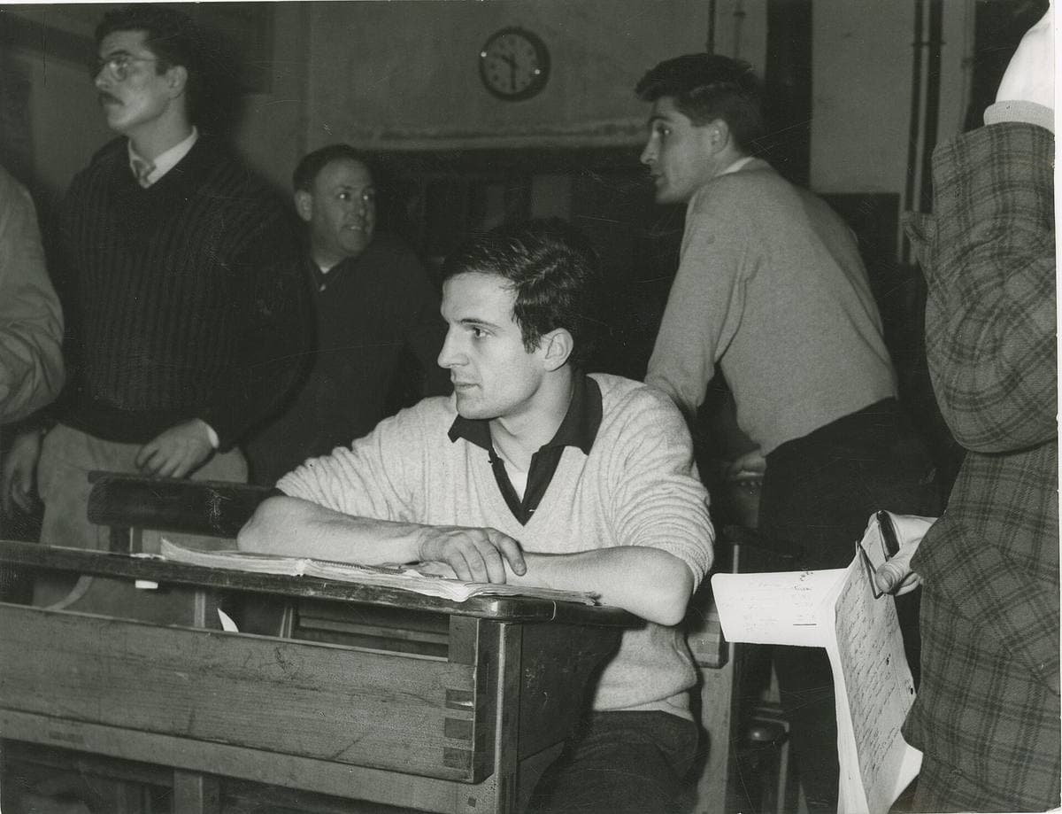François Truffaut in a pale sweater, leaning on an old wooden school desk with a book open in front of him