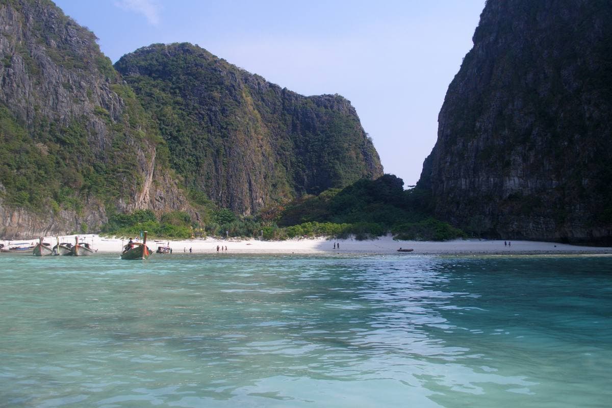 A white beach under limestone cliffs, with people on the sand and boats at the shore