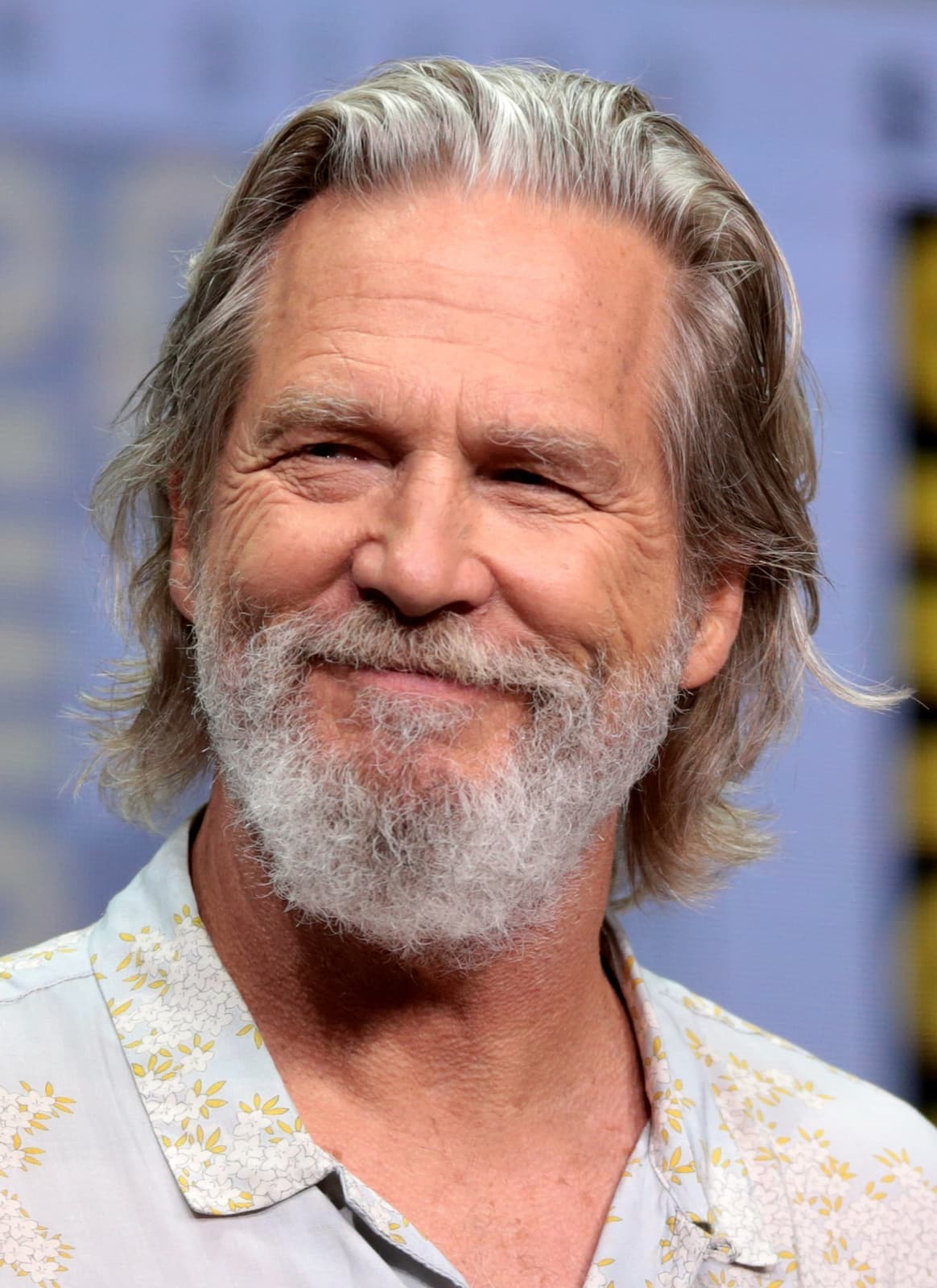 Jeff Bridges smiling, grey hair and beard, in a light patterned shirt
