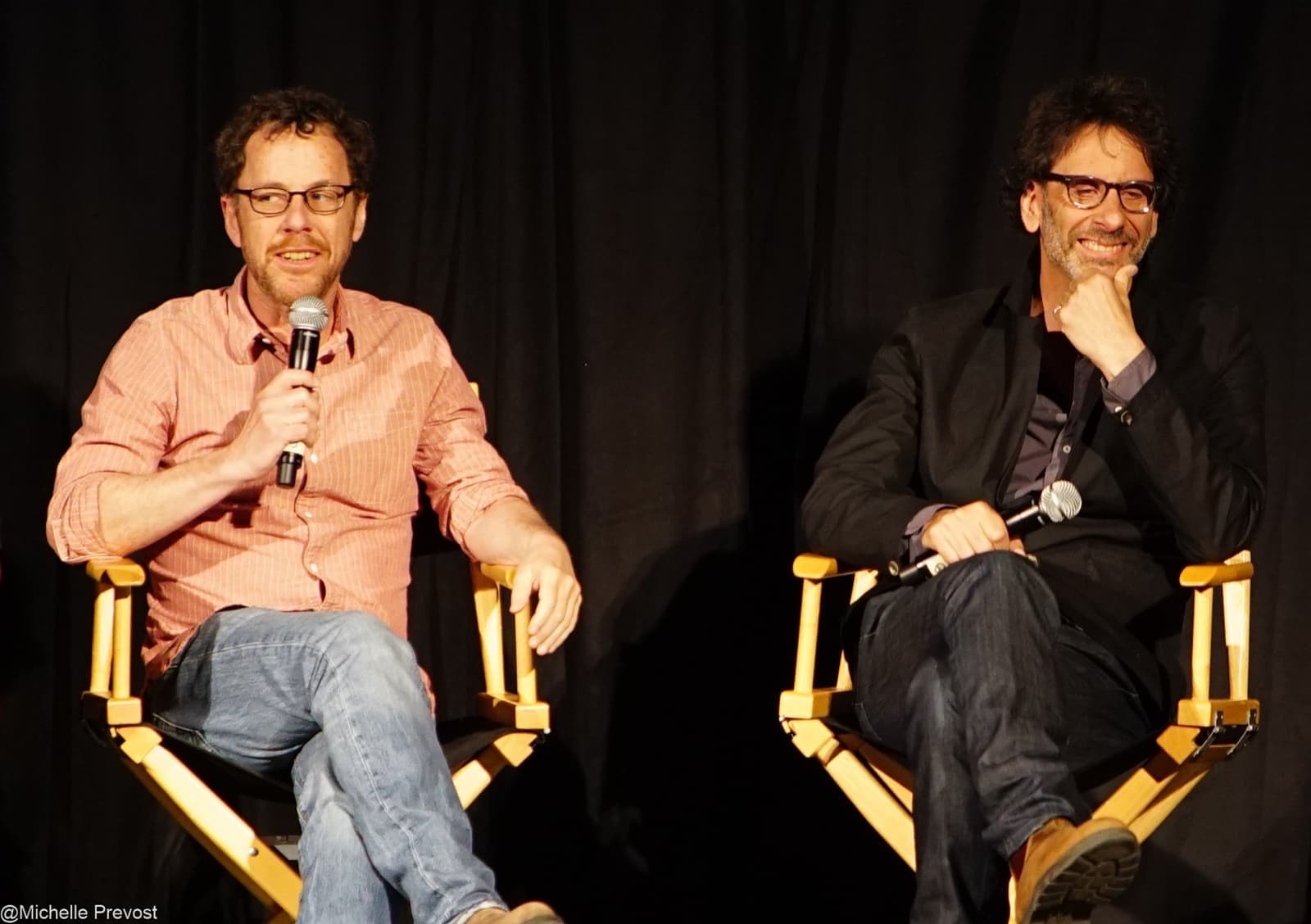 Ethan Coen and Joel Coen seated in director’s chairs on a dark stage