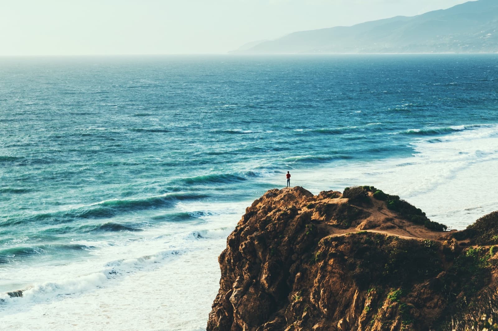 A figure standing on the rocky tip of Point Dume above blue surf
