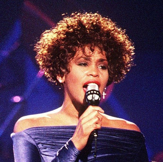 Whitney Houston singing into a microphone in an off-the-shoulder blue gown