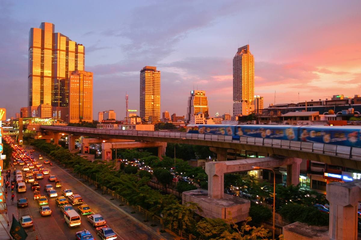 A city avenue at dusk with traffic, an elevated railway and tall towers