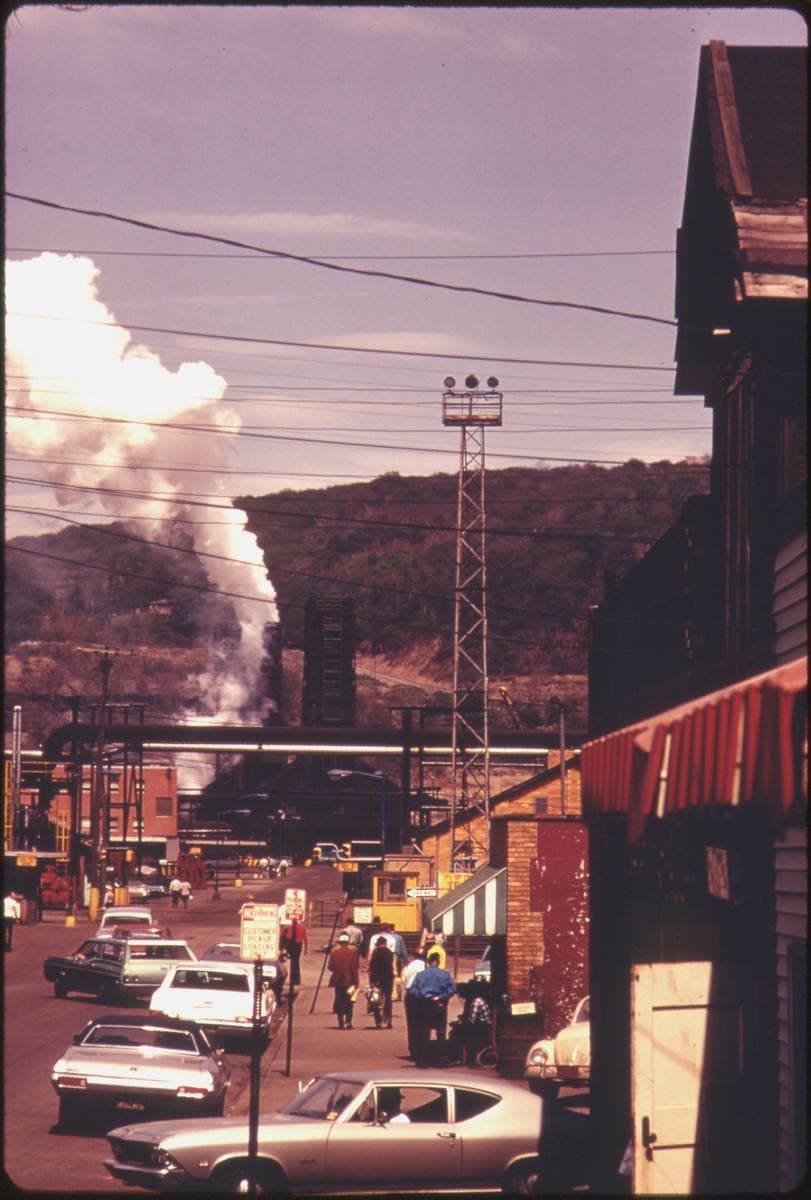 A steep street of shops and parked 1970s cars running down towards a smoking industrial plant