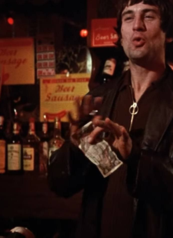 A young Robert De Niro in a dark shirt in a bar, holding a banknote