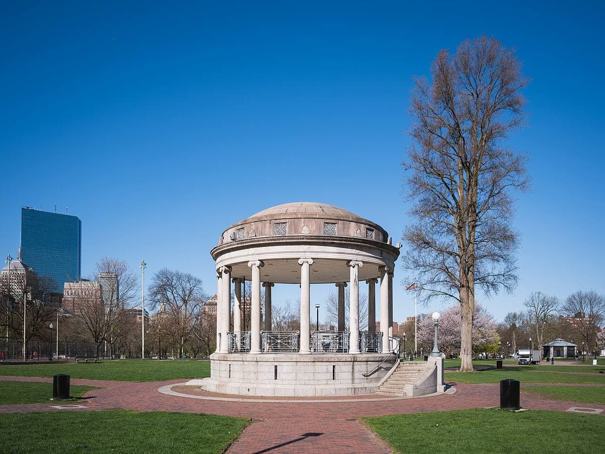A round white bandstand with columns on a lawn, tall buildings beyond