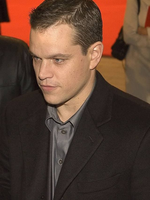 Matt Damon in a dark coat and grey shirt, looking down