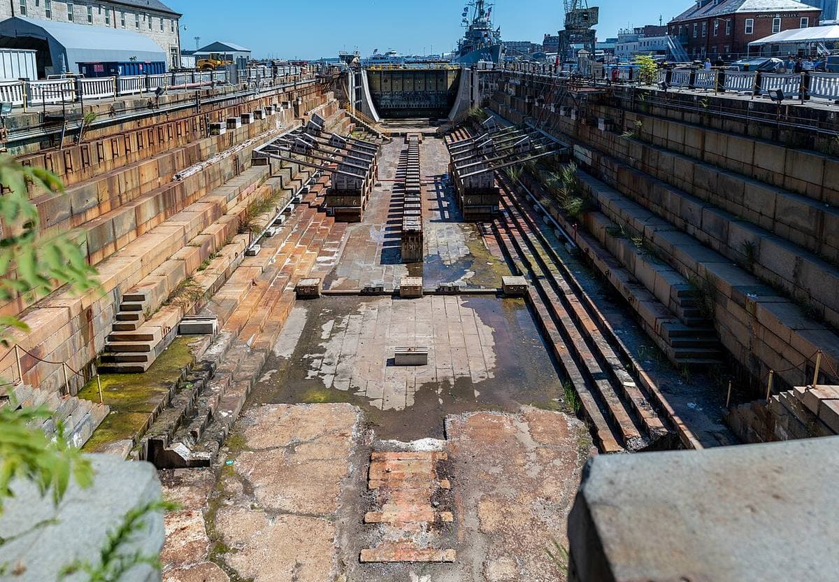 A granite dry dock with a ship in it, harbour buildings beyond