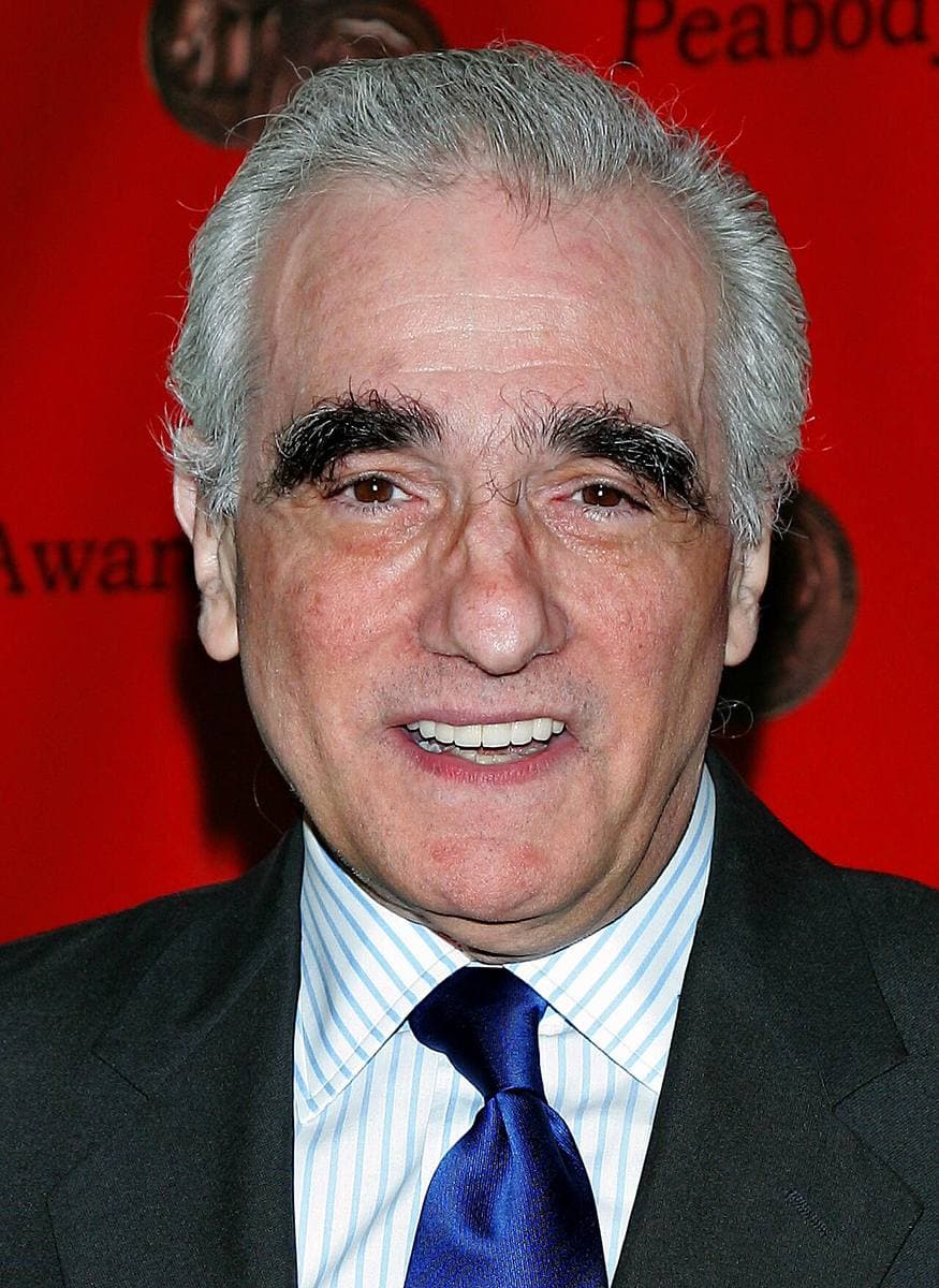 Martin Scorsese in a blue tie, smiling, against a red backdrop