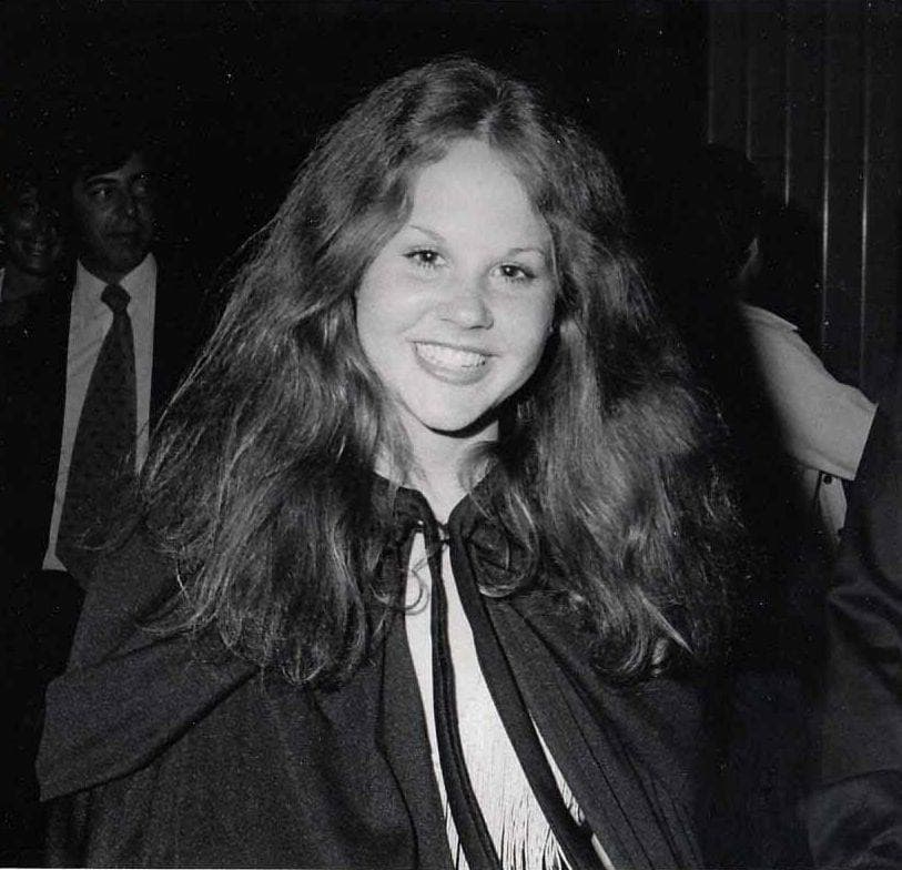 Linda Blair in 1976, long hair, smiling, in a dark cape