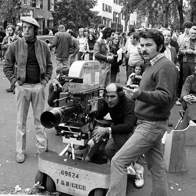 Black-and-white photograph: a man in a flat cap and glasses stands by a film camera on a dolly in a leafy Georgetown street, with a crowd of onlookers behind