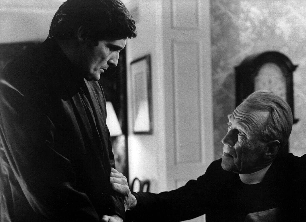 Two priests in black, the younger standing, the older looking up at him