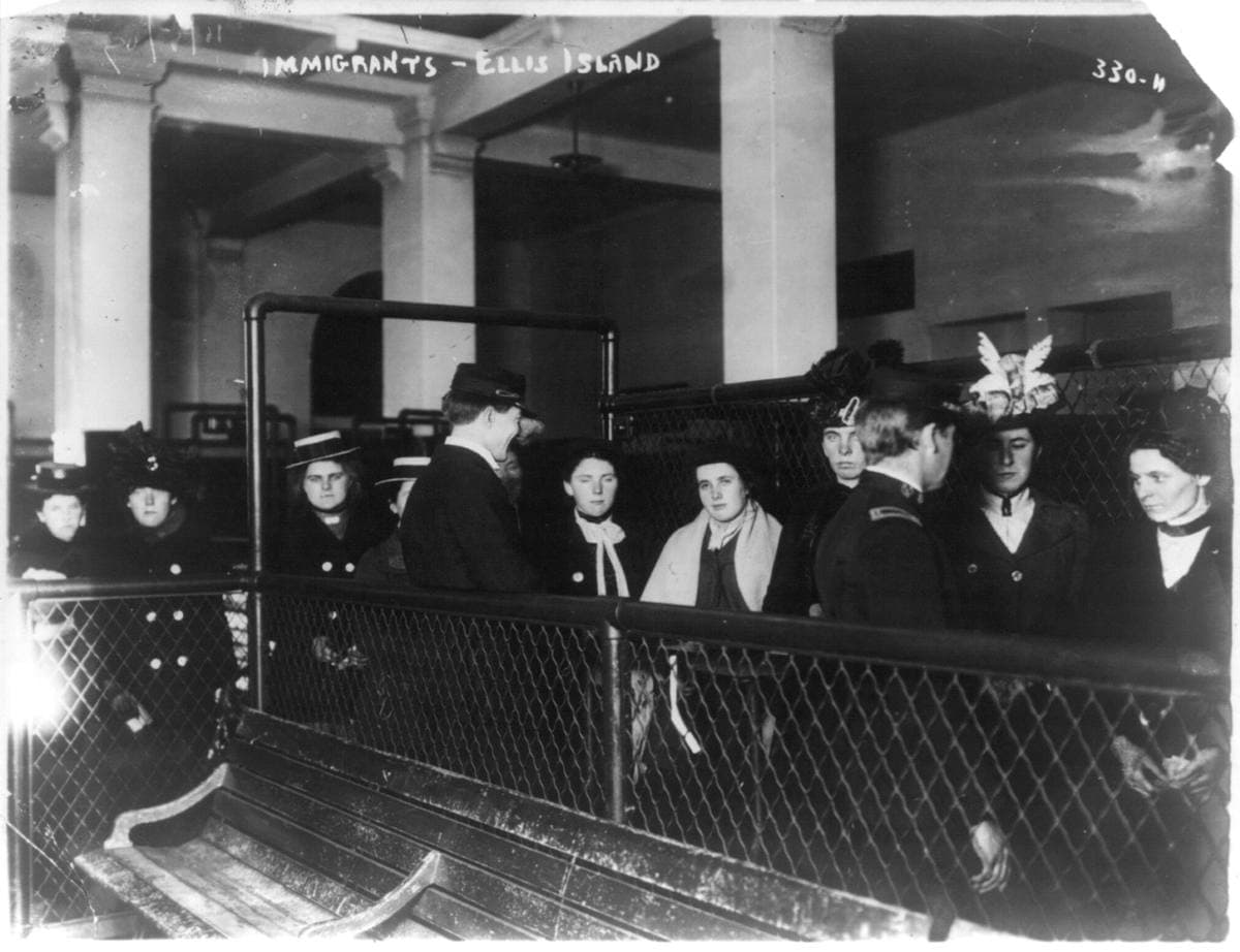 Women in hats and shawls waiting behind iron railings in a hall at Ellis Island