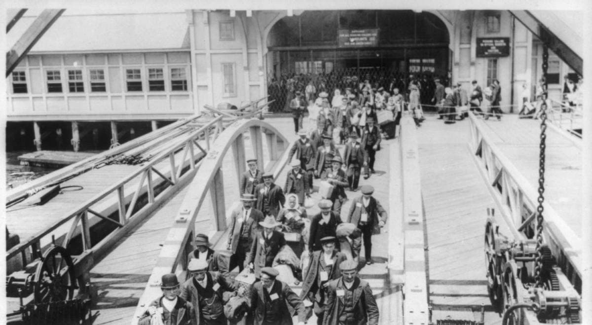 A crowd of new arrivals in coats and hats walking up a covered gangway towards the Ellis Island building