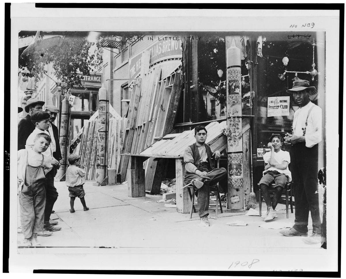 Men and boys around a street stand hung with decorations in Little Italy, with tenements behind