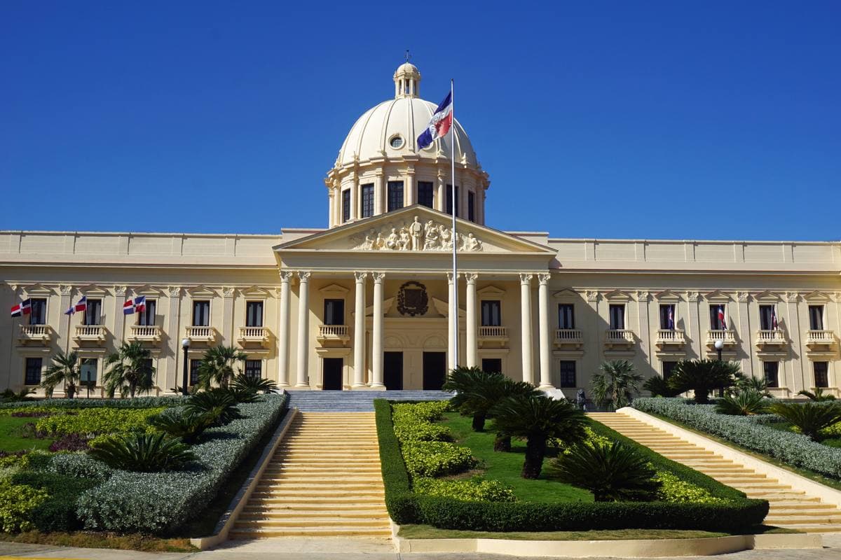 A pale neoclassical palace with a columned portico and a dome behind railings and palms