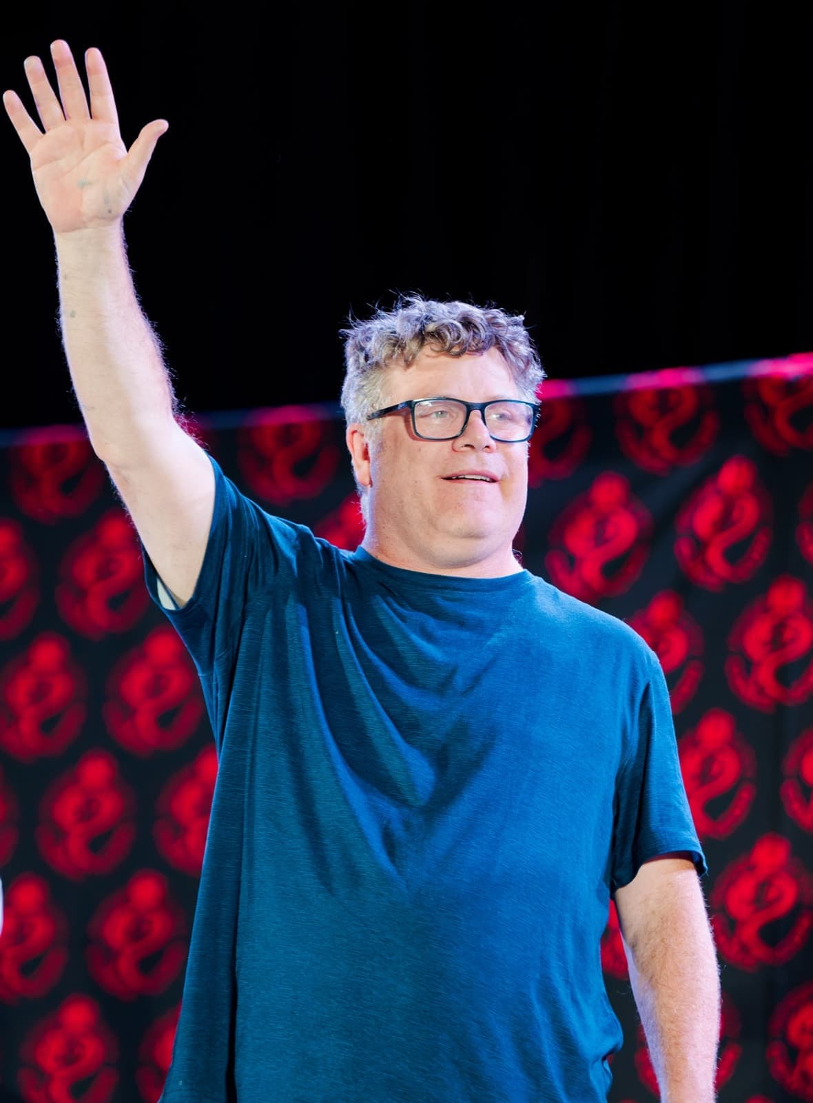 Sean Astin on a stage, waving with one arm raised high, grinning, in glasses and a blue T-shirt