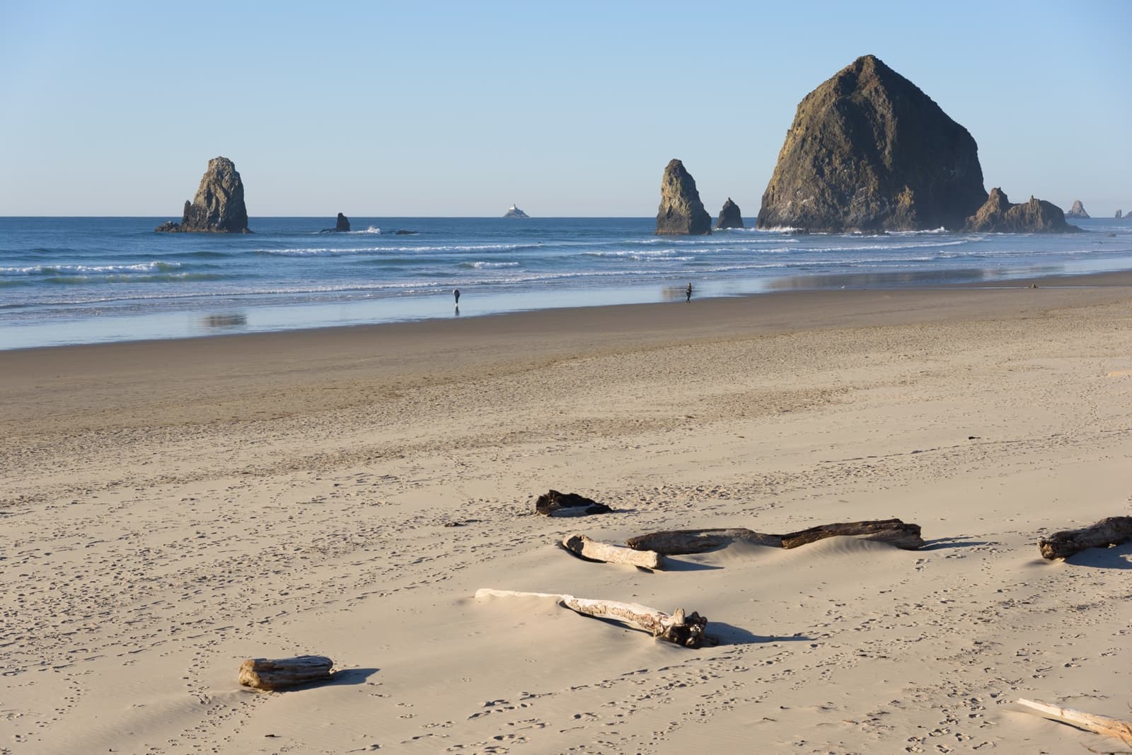 A wide sunny beach with driftwood in the foreground and Haystack Rock and the Needles offshore