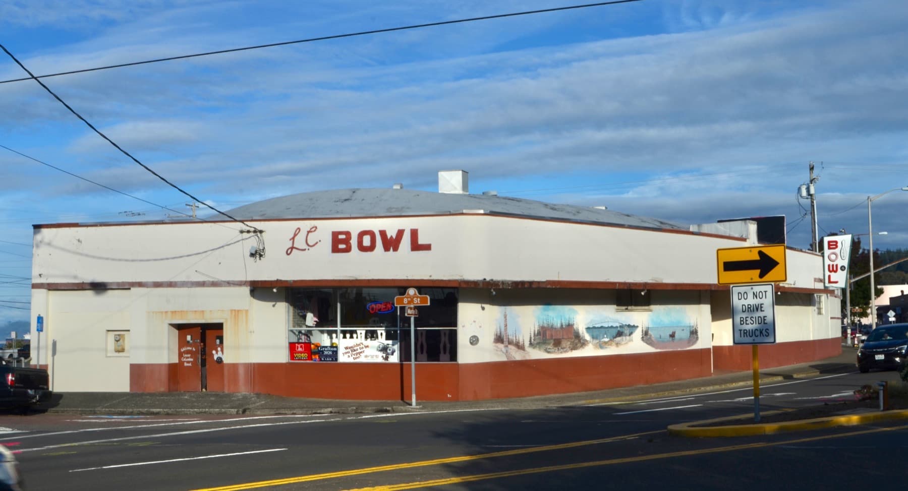 A low white bowling alley with a red Bowl sign and painted murals along its side, on a sunny street corner