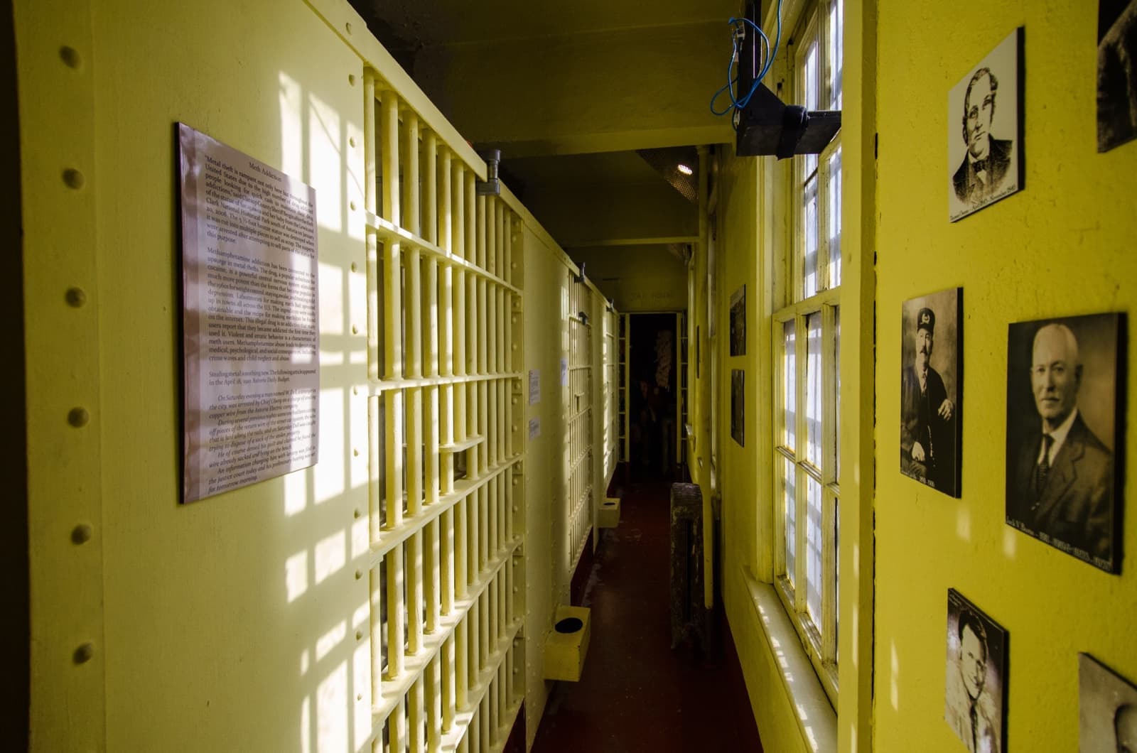 A narrow yellow corridor beside barred cells, framed photographs on the wall