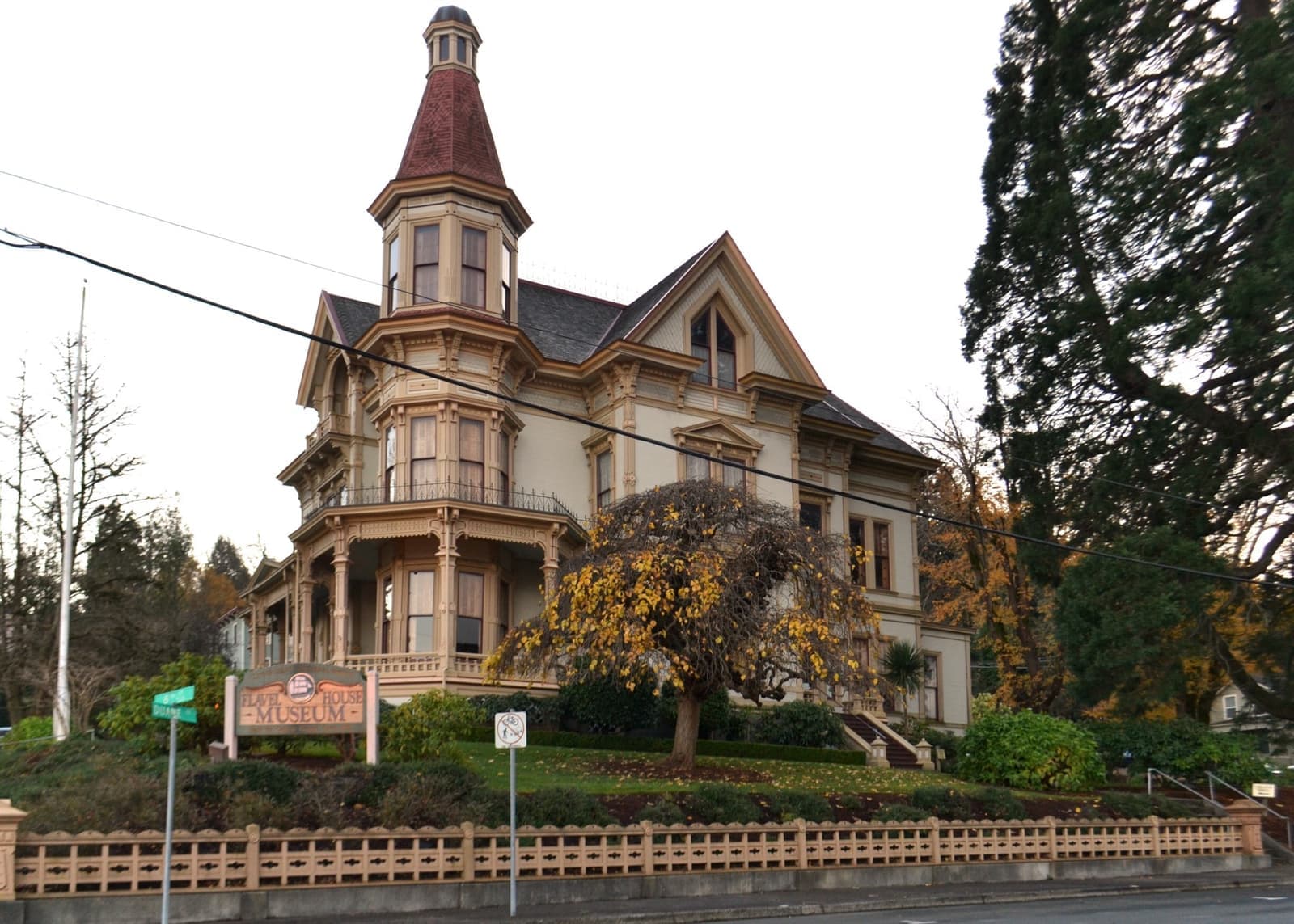 A tall Queen Anne house with a pointed corner tower, verandas and a picket fence