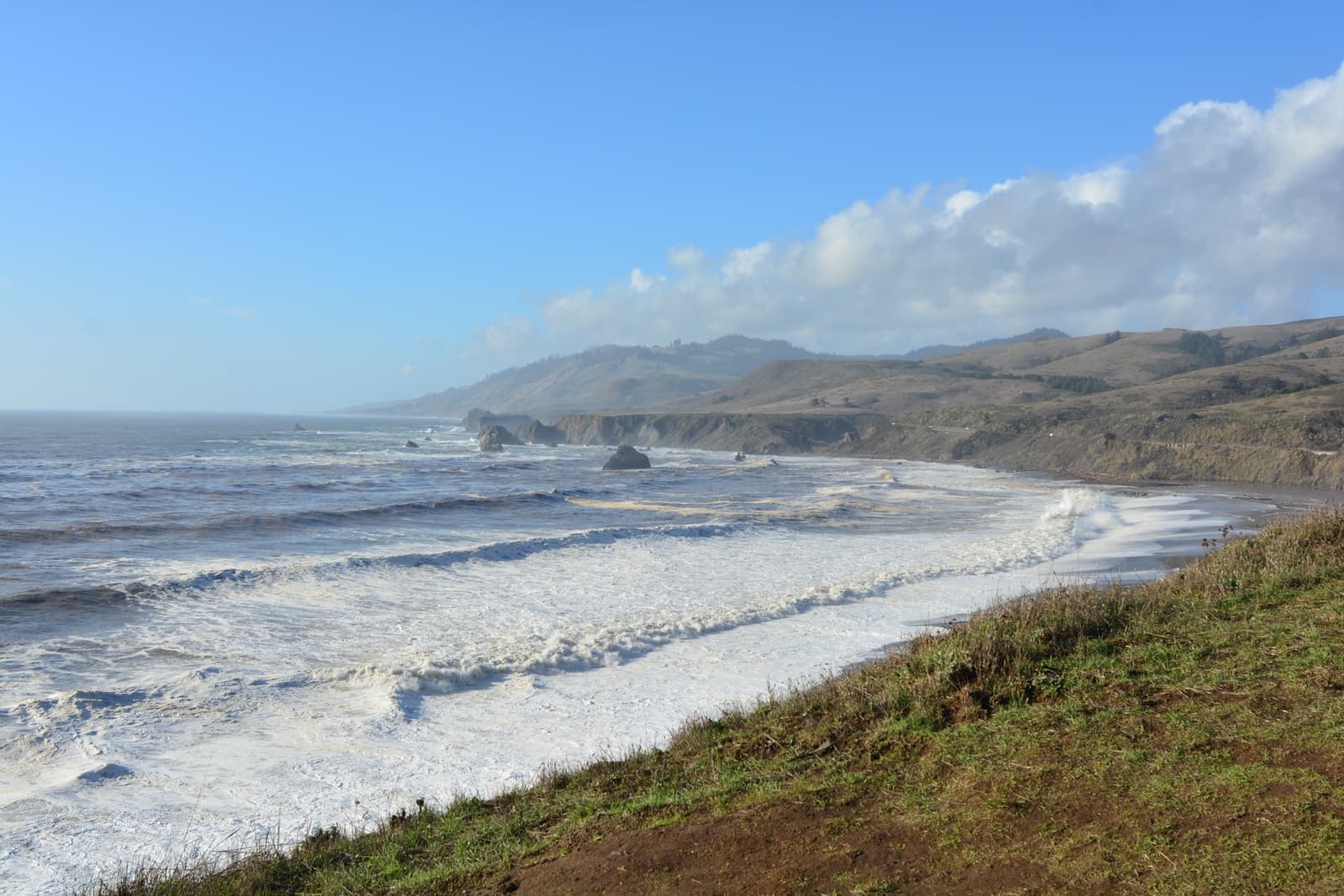 A long sandy spit and breaking surf below green bluffs, sea stacks offshore