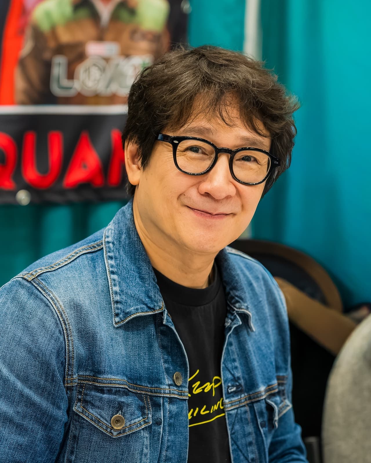 Ke Huy Quan smiling in round black glasses and a denim jacket
