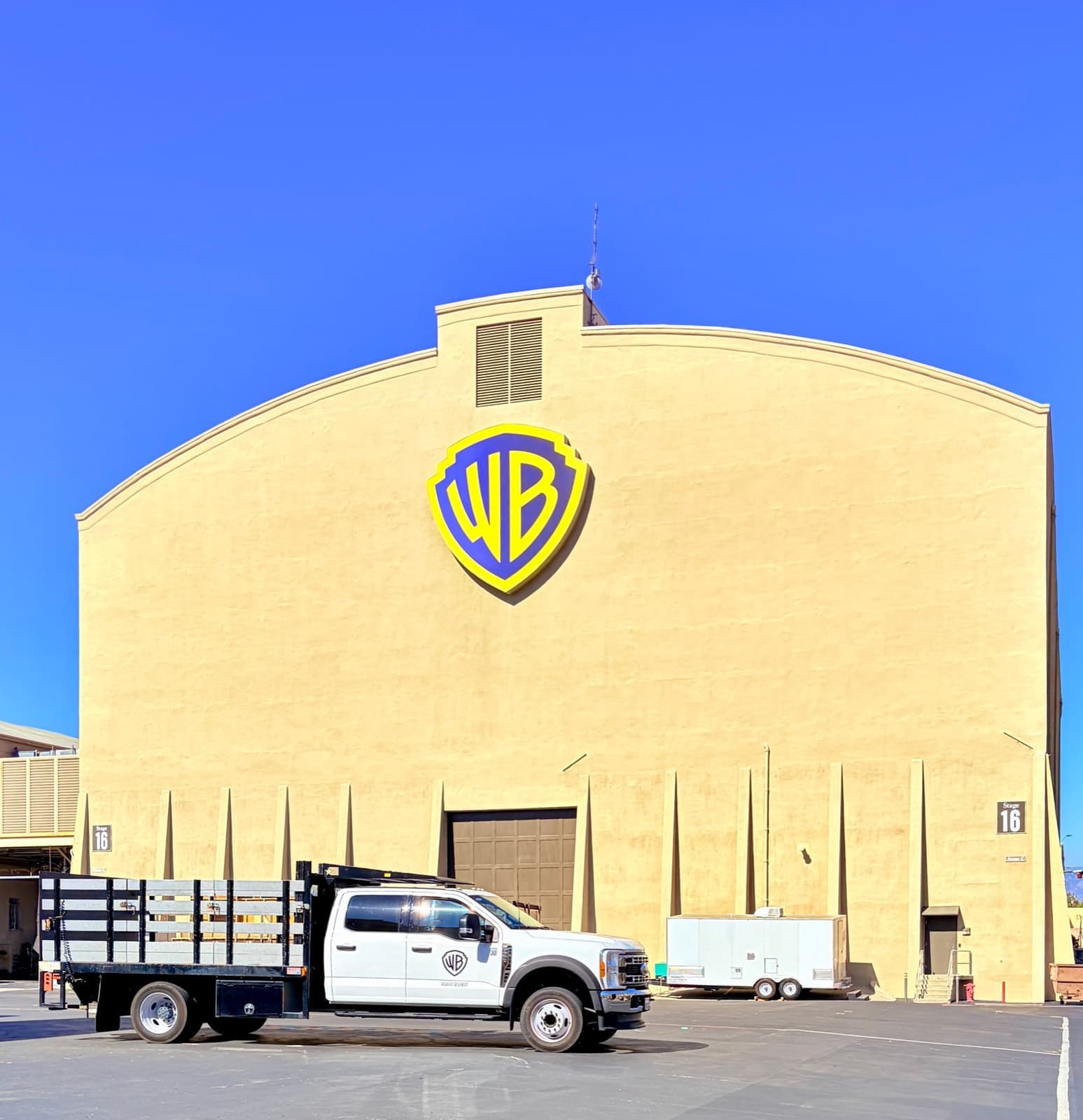 A tall cream sound stage with a big studio shield on its gable and a truck parked below