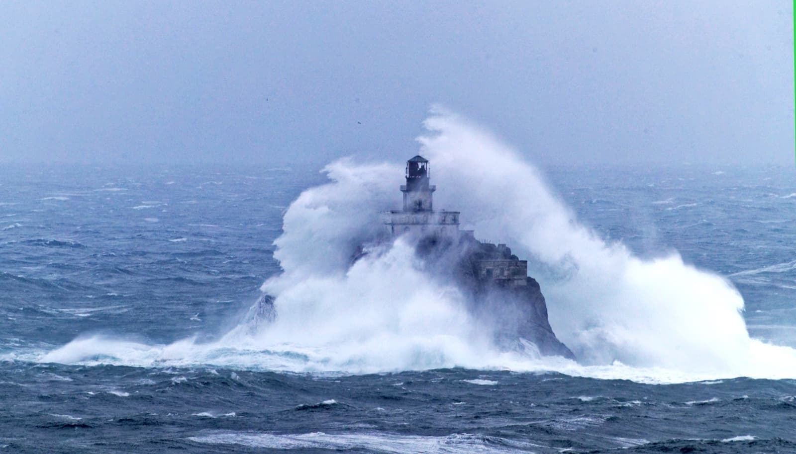 A wave bursting white over a small lighthouse on an offshore rock