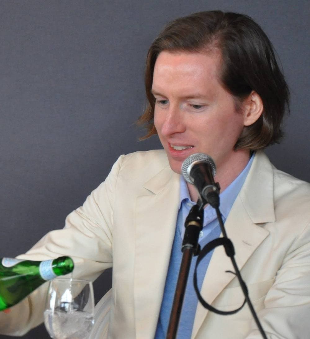 Wes Anderson in a cream suit pouring water at a press table