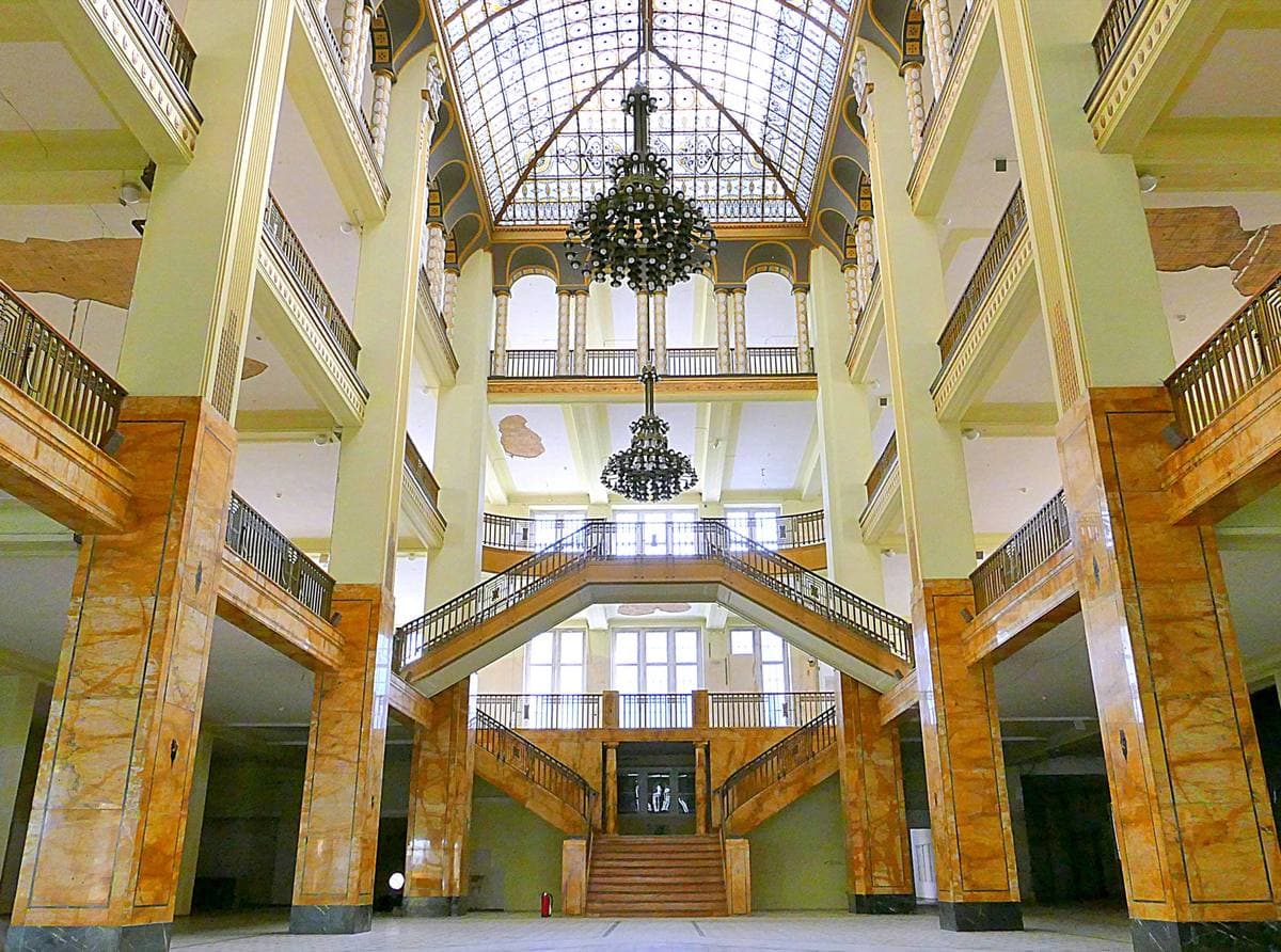 A tall atrium with marble-clad pillars, gallery balconies, two chandeliers and a double staircase under a glass roof
