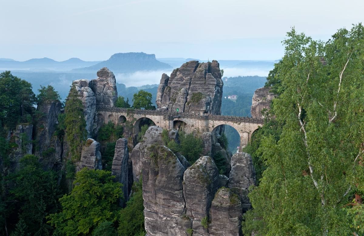A stone bridge of arches linking sandstone towers above forest, mist in the valley beyond