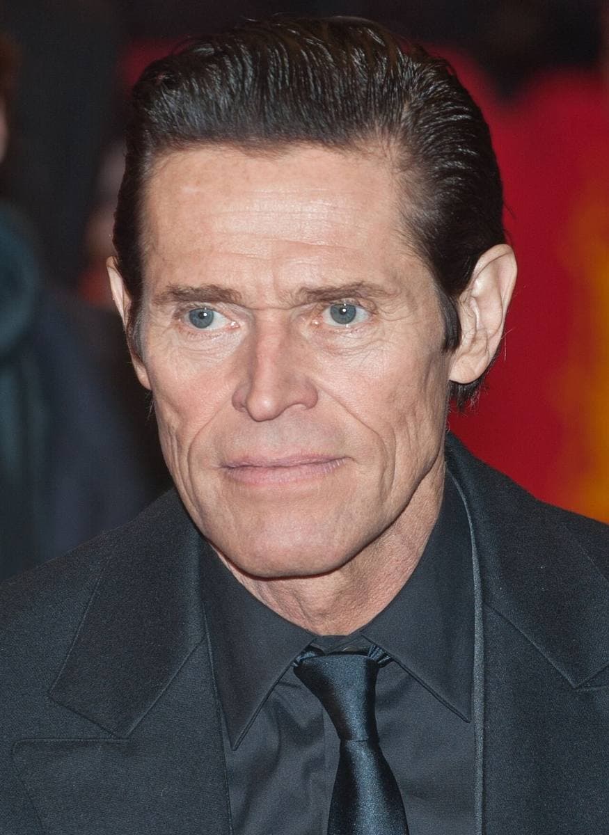 Willem Dafoe in a black shirt and tie against a red backdrop