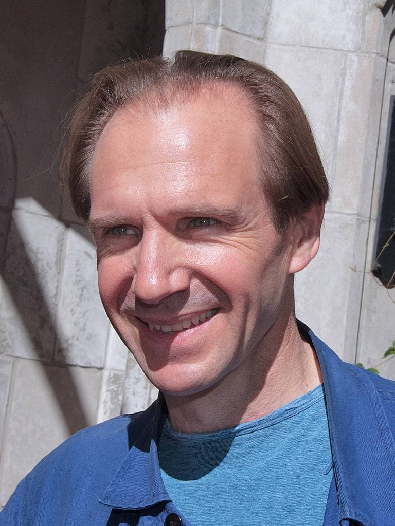 Ralph Fiennes smiling in a blue shirt against a pale stone wall