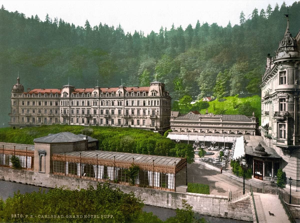 A hand-coloured view of a long hotel in a wooded valley, with gardens and a river