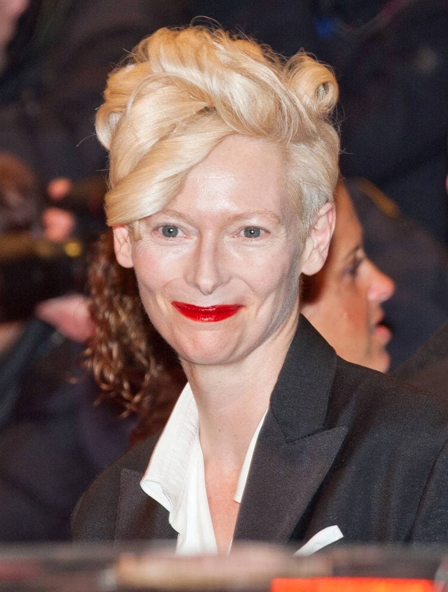 Tilda Swinton with swept-up blonde hair and red lipstick on a crowded red carpet