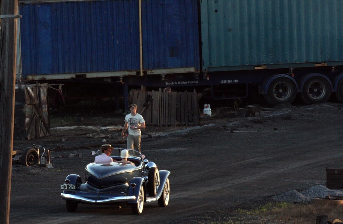 A vintage open-top car driving across a gravel yard in front of stacked shipping containers, a man in a T-shirt walking behind it
