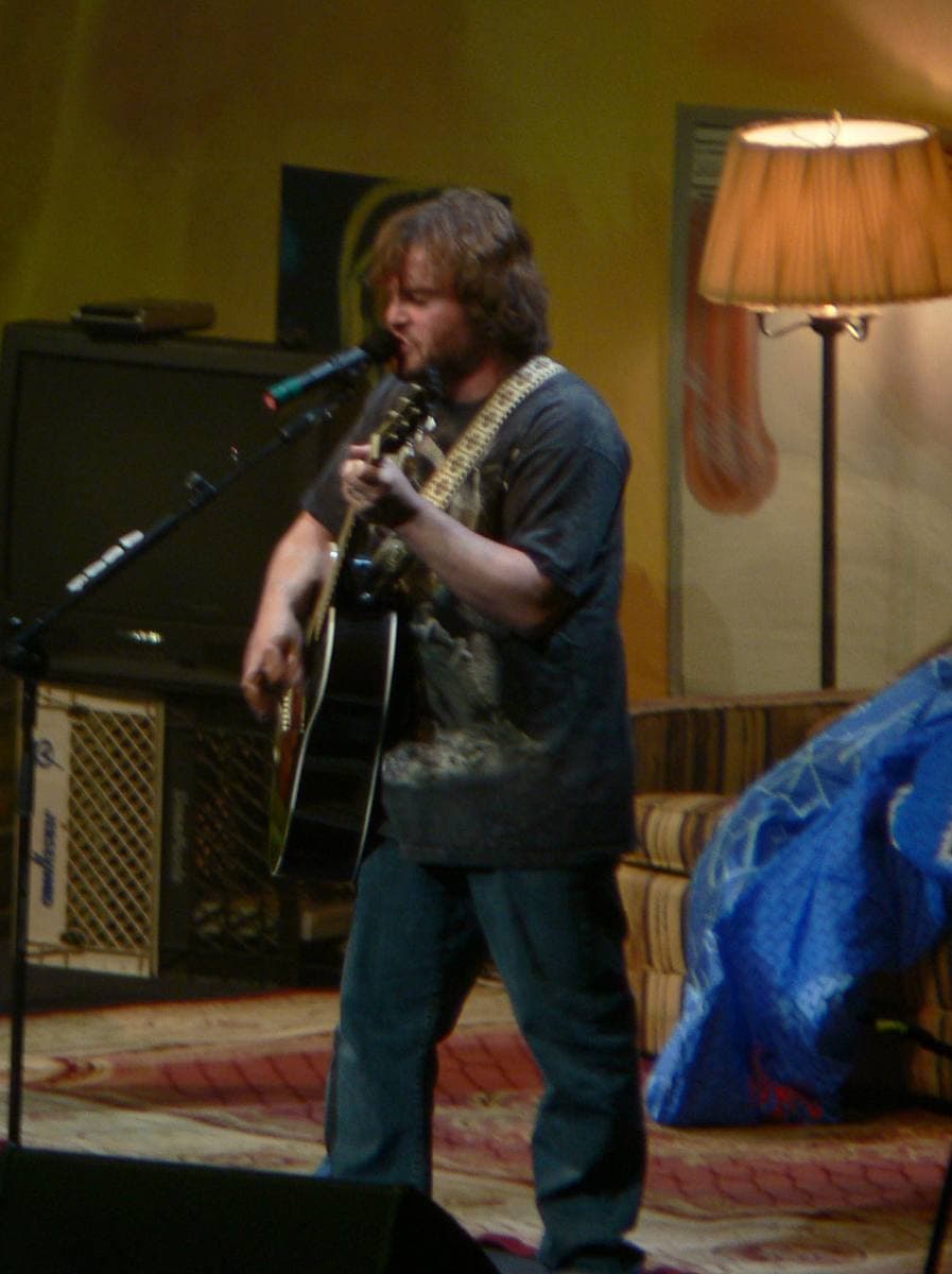 Jack Black singing into a microphone and playing an acoustic guitar beside a lamp on stage