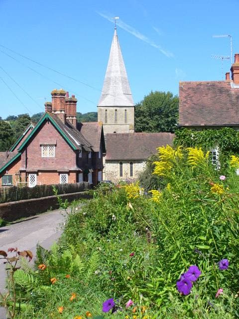 A lane of tile-hung brick cottages running downhill to a church spire, with yellow and purple flowers in front