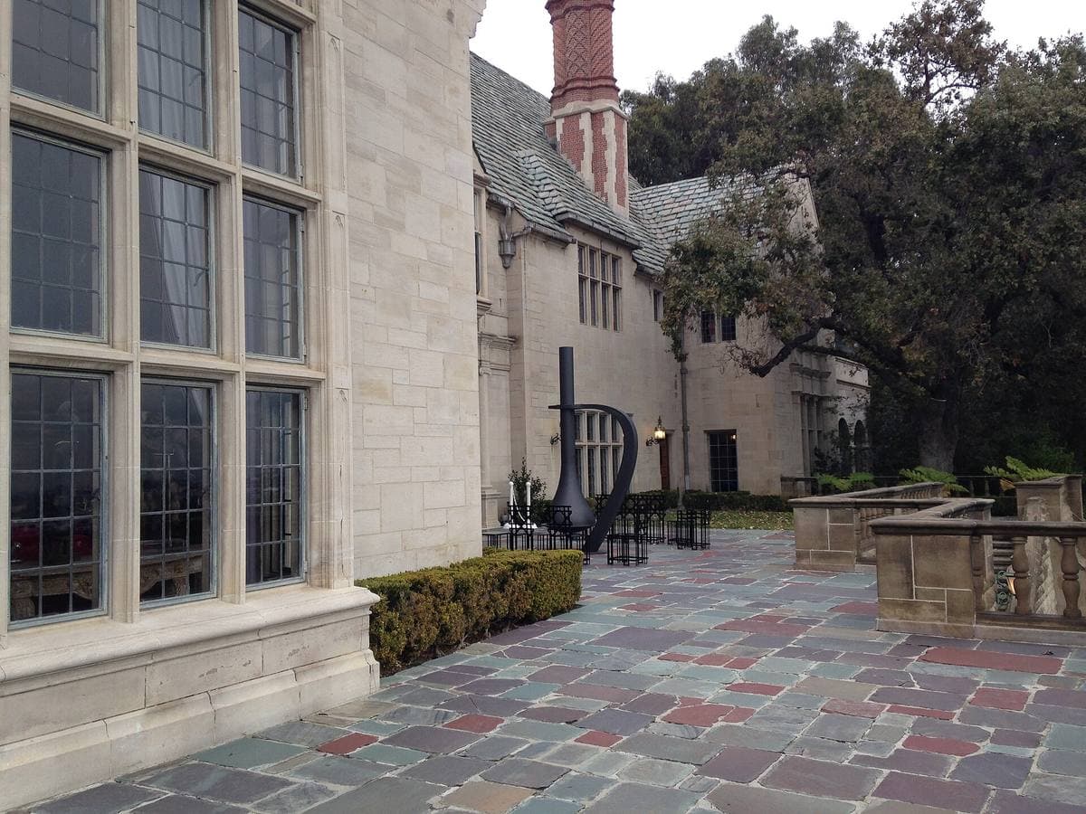 A grey stone Tudor-style mansion with tall leaded windows above a paved terrace