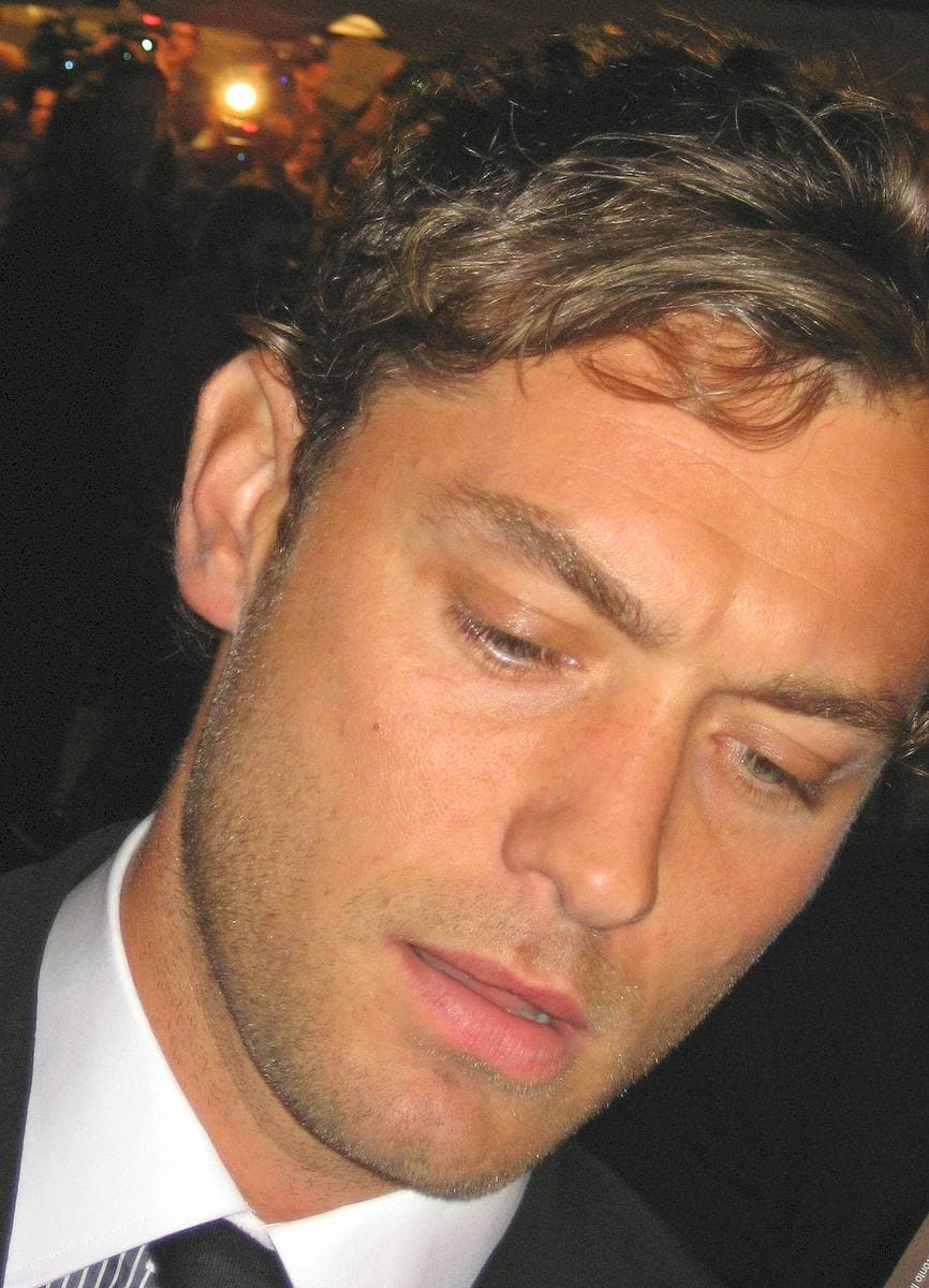 Jude Law in close-up, in a dark suit and tie, looking down