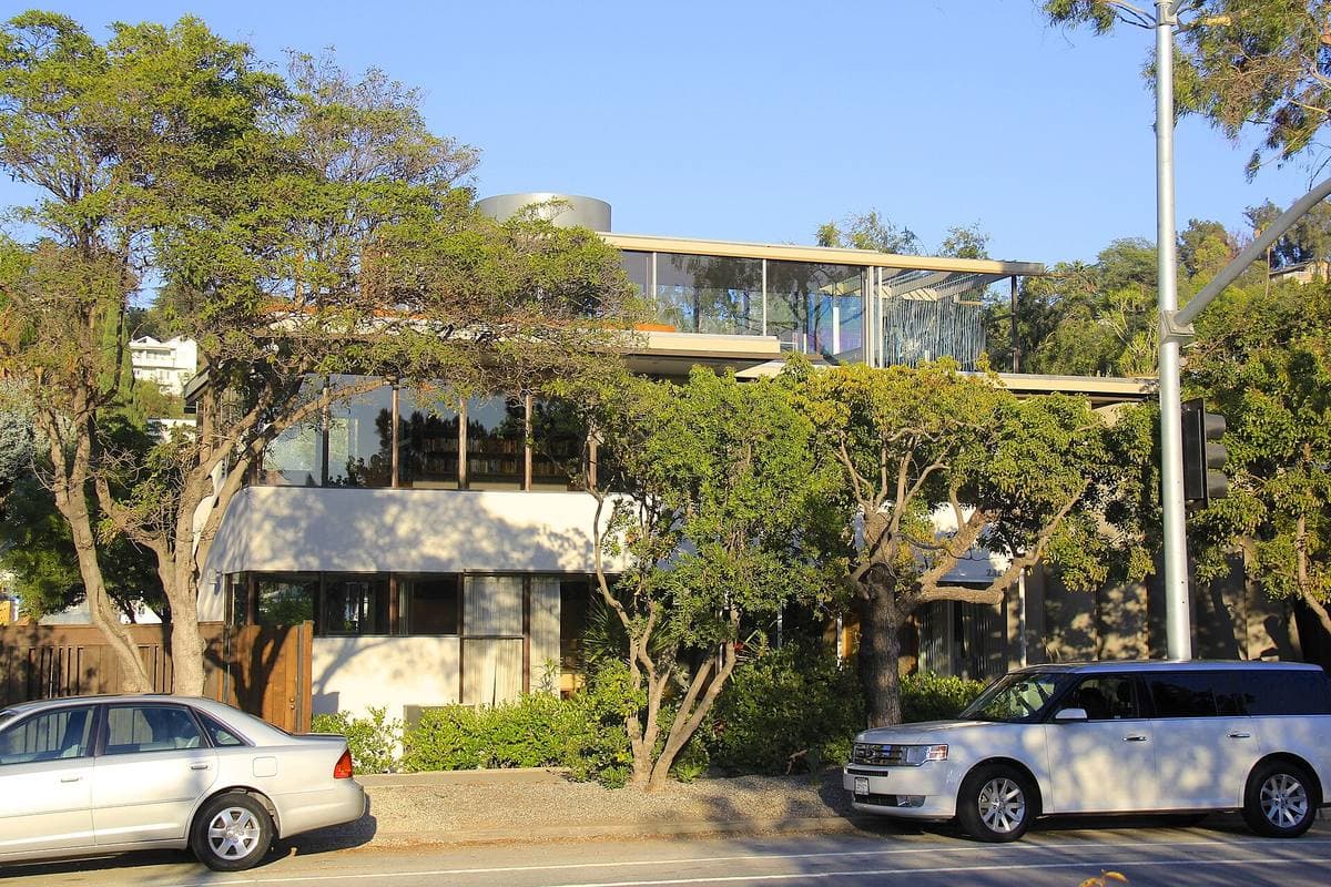 A low glass-and-white modernist house among trees by a road