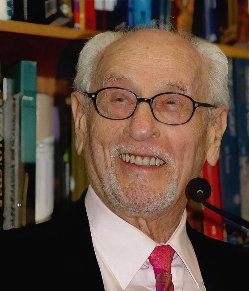 Eli Wallach, white-haired and smiling broadly in glasses and a red tie, in front of bookshelves