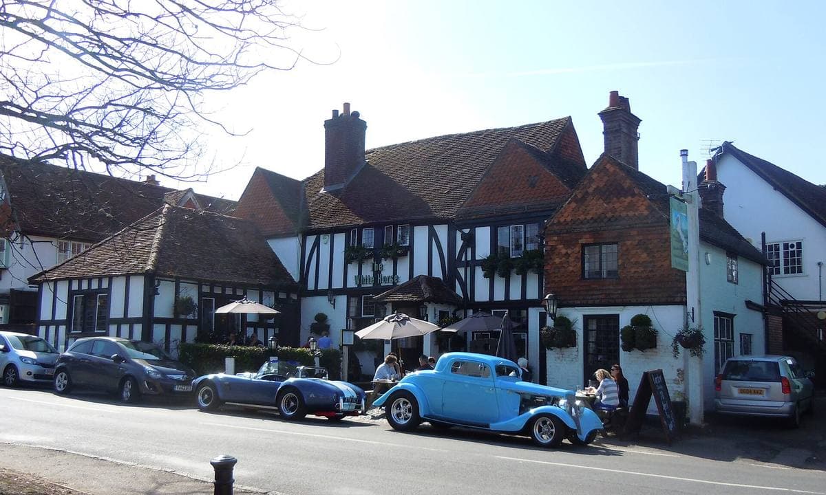 A long timber-framed pub with white plaster and tile-hung gables, old cars parked outside