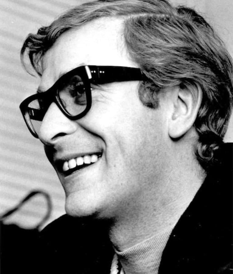 Black-and-white photograph of Michael Caine in heavy black glasses, laughing