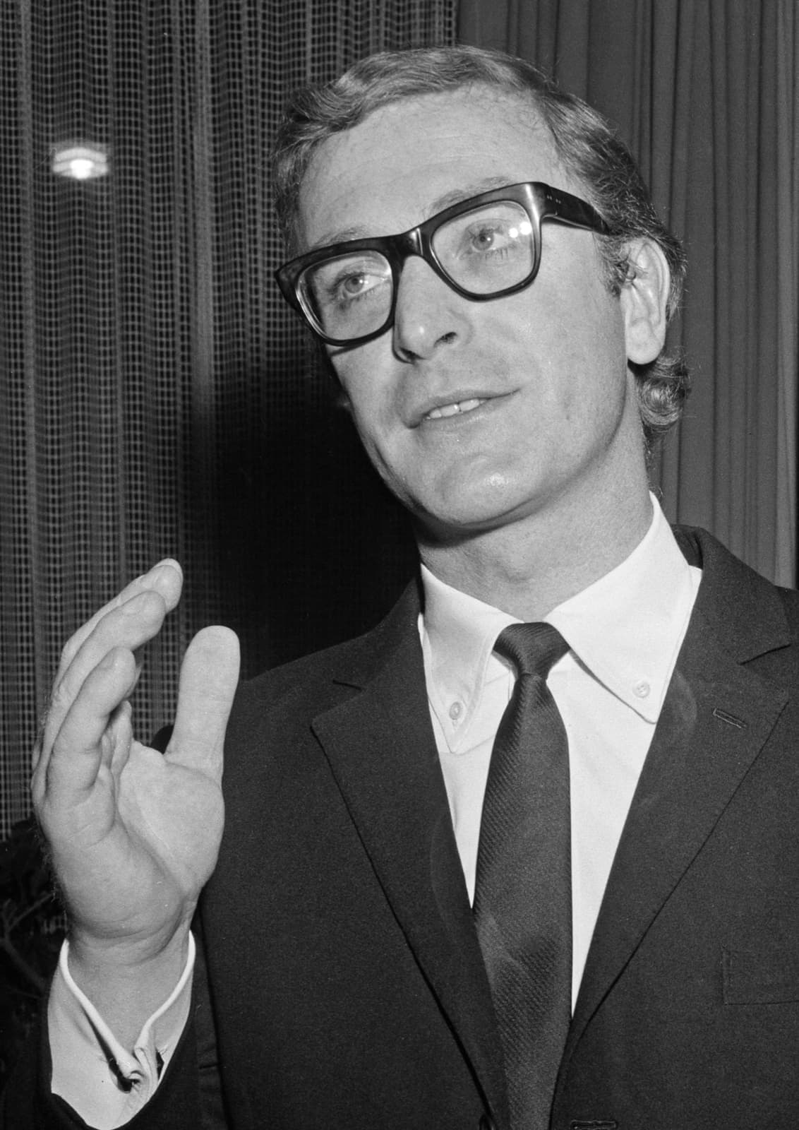 Black-and-white photograph of Michael Caine in a dark suit and glasses, one hand raised as he talks
